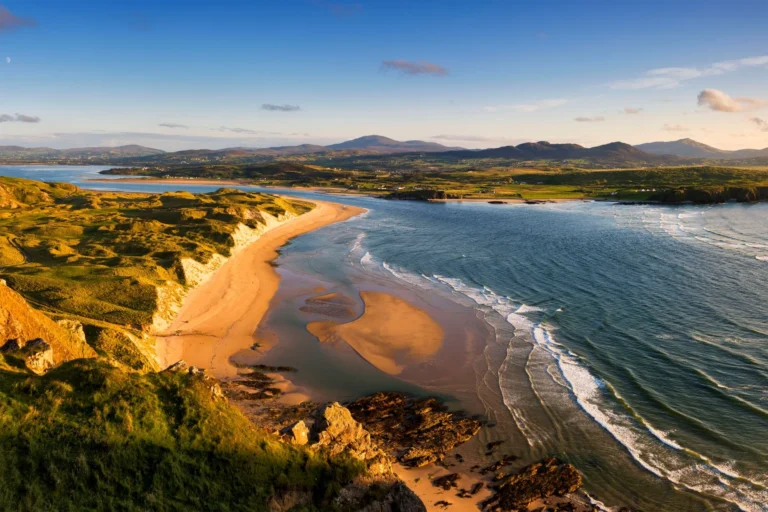 Panoramic view of Inishowen Peninsula coastline with sandy beach, rolling dunes, Atlantic waves and distant green hills under clear sky